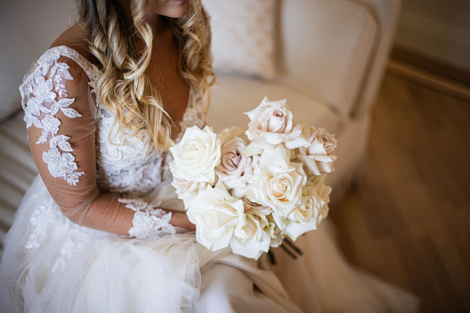 Foto di una sposa, seduta su un divano chiaro, che tiene in mano un elegante bouquet di rose bianche, pronta per andare in chiesa dove tutto è allestito per il suo matrimonio.