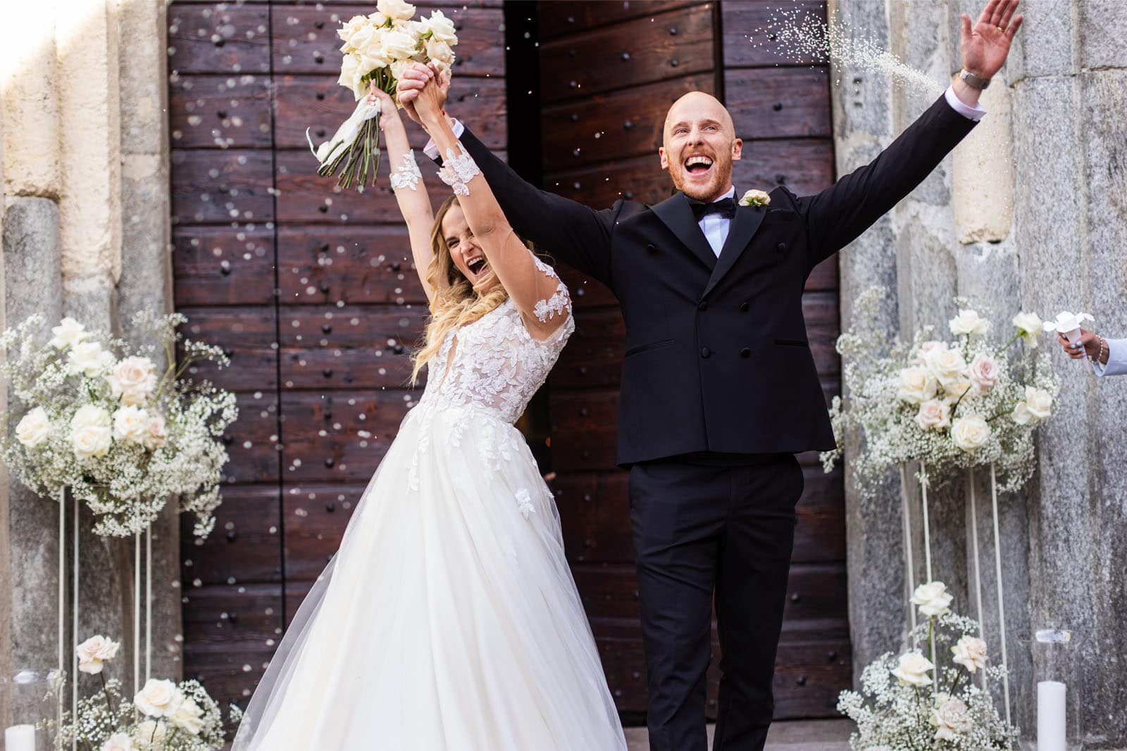 Coppia di sposi felici, appena usciti dalla chiesa, che esultano a braccia aperte sollevando al cielo un bouquet di rose bianche. Alle loro spalle si vedono degli elementi floreali, formati da rose bianche come il bouquet.