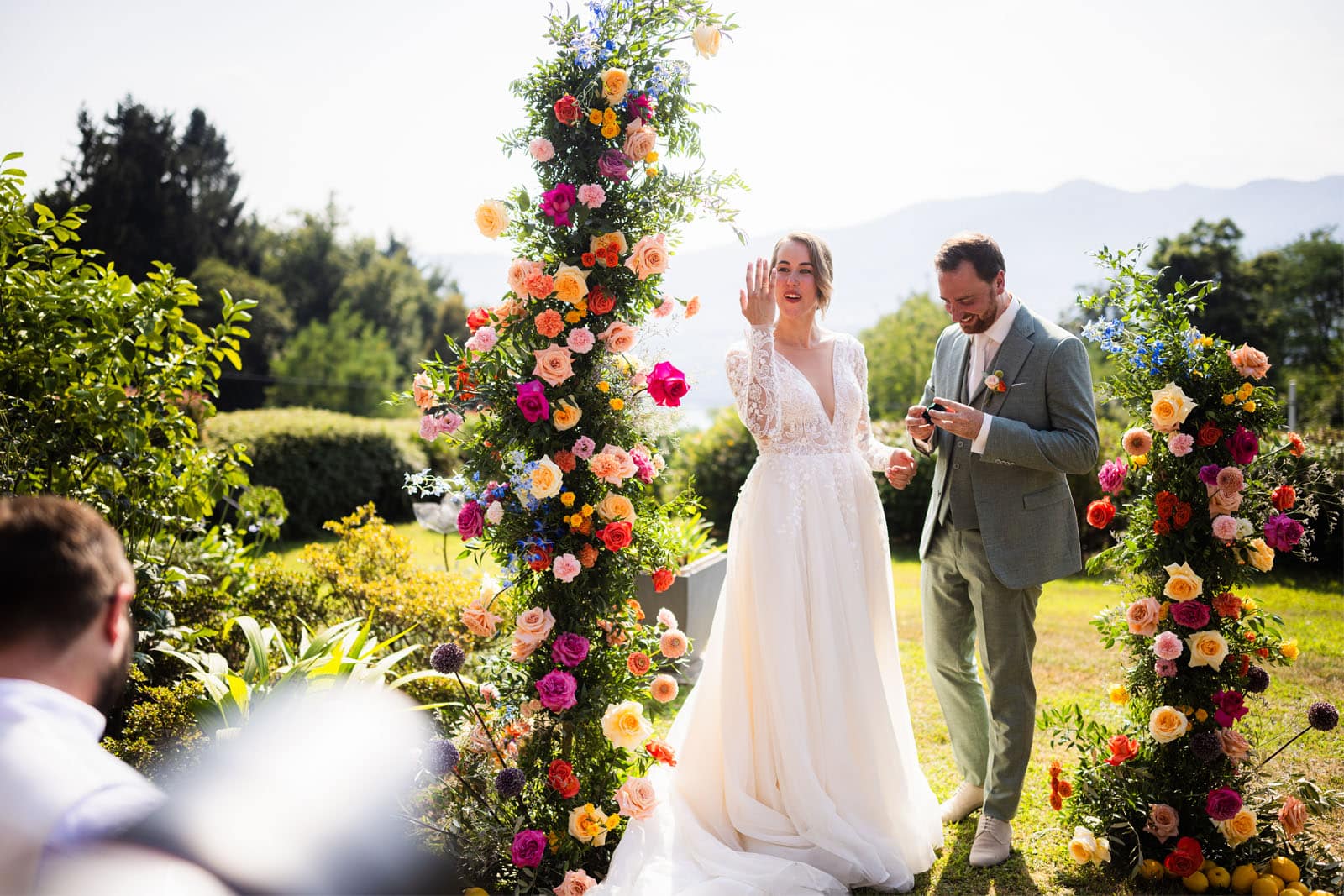 Coppia di sposi, situati all'interno di un arco di fiori colorati, allestito all'aperto. La foto, scattata durante la parte finale del matrimonio, si concentra sull'espressione felice della sposa intenta ad ammirare, felice, la sua fede nuziale.