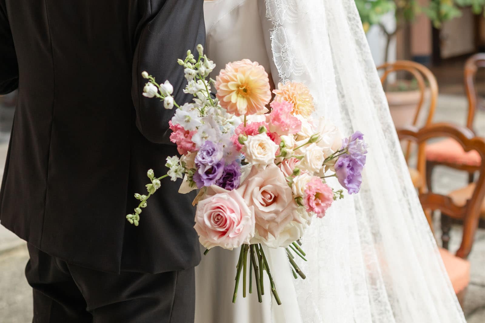 Primo piano di un bouquet sorretto da una sposa. Esso è composto da fiori di vario tipo e dimensione, e la prevalenza di colori usati sono quelli pastello tra le gradazioni di rosa e lilla.