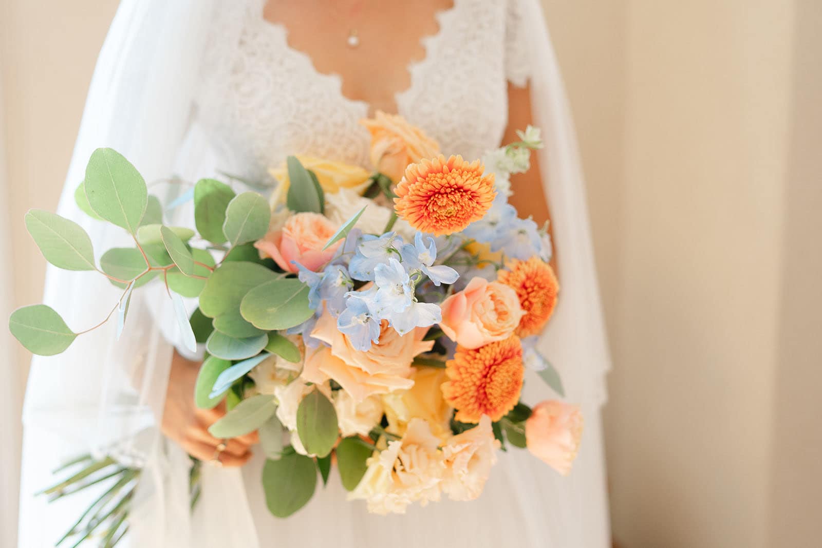 Primo piano di un bouquet, sorretto da una sposa, composto da fiori di colore caldo e dalle sfumature arancioni. A dare contrasto all'insieme, nella parte centrale sono presenti alcuni fiori di un tenue color azzurro.